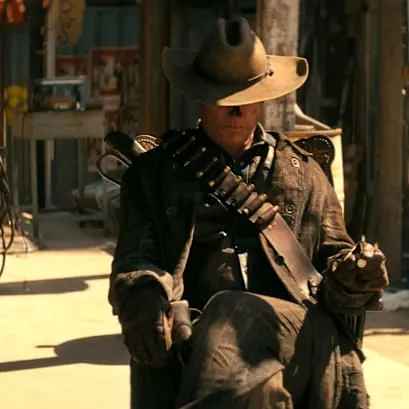 The Ghoul sits casually with his hat tipped over his eyes in the center of a cobbled-together town